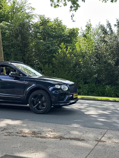 A black Bentley is parked on an asphalt road next to a grassy verge, with lush green trees in the background. The location is Dronten, Netherlands. The car's front is visible, showcasing the headlights and the black rims with the Bentley logo. The road surface shows some markings and debris. Sunlight filters through the trees, casting shadows on the road.