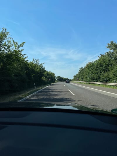 The image is taken from inside a vehicle, looking out at a highway. The perspective is angled, as if the camera is tilted or the photo was taken while moving. The scene depicts a bright, sunny day with a clear blue sky dotted with wispy white clouds. On either side of the road, lush green trees form a dense border.
The highway itself appears to be asphalt with a white line marking the lanes. There is a white and orange striped barrier on the left side of the road. In the distance, a dark-colored car is visible driving in the same direction. Along the right side of the road, a series of rounded reflectors or markers are visible. There is also a triangular warning sign with a red border and a black symbol (possibly depicting a curve or hazard) visible amongst the trees on the left.
The reflections on the side of the vehicle from which the photo is taken suggest it is a dark-colored car. The overall impression is one of a scenic drive on a pleasant day. The location is identified as Sankt Augustin, Germany. No people are clearly visible, and the focus is on the landscape and the road.
