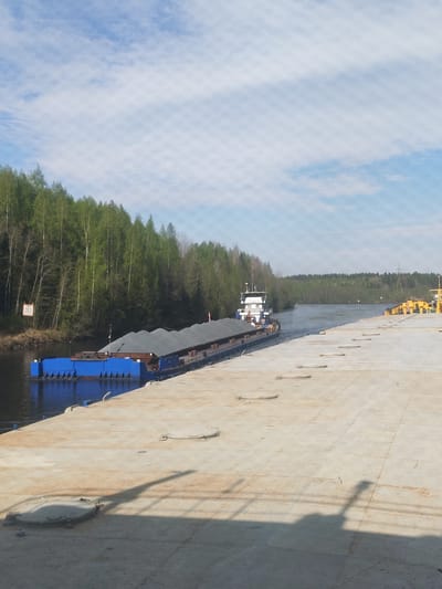 The image shows a barge loaded with gray material, possibly gravel, on a waterway in Depo, Russia. A tugboat is pushing the barge. A concrete pier is visible in the foreground, with equipment on the far side. The waterway is lined with trees, and the sky is partly cloudy.