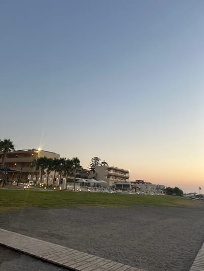 The image depicts a seaside resort area in Rhodes, Greece, likely during the late afternoon or early evening as indicated by the soft, warm light of the setting sun. The sky transitions from a clear, pale blue on the left to a gradient of orange and yellow towards the horizon on the right.
The scene is dominated by a long, paved walkway bordering a strip of green grass. Beyond the grass, a multi-story resort building is visible, characterized by white or light-colored facades, numerous balconies, and rows of windows. Palm trees are interspersed along the front of the resort, adding to the tropical ambiance. String lights are visible along the resort's exterior, suggesting it is preparing for evening illumination.
In the background, other resort buildings and some trees can be seen. A flagpole with a Greek flag is also present, reinforcing the location. The ground in the foreground appears to be a mixture of sand and gravel, typical of a beach environment. There are no people visible in the frame. The overall mood is peaceful and serene, capturing the tranquil atmosphere of a coastal destination at dusk.