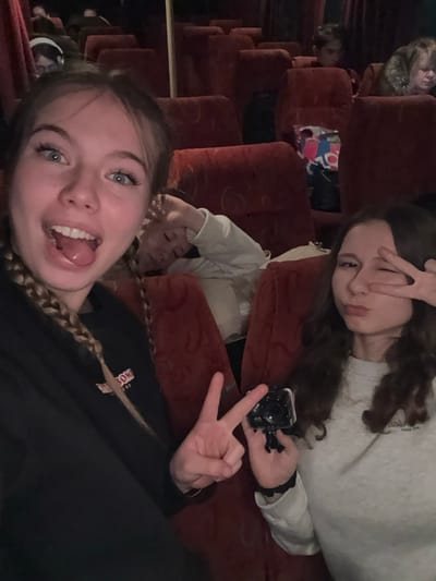 An image taken inside a passenger vehicle, likely a bus or coach, in Chernovskoe, Russia, shows multiple individuals seated in rows of red, upholstered seats under dim lighting conditions.
In the foreground, two young females are posing for the camera. The female on the left has light-colored hair styled in braids, prominent blue eyes, and is wearing a dark top. She displays an open-mouthed expression with her tongue slightly extended and makes a "peace sign" gesture with her left hand. The female on the right has dark, wavy hair and is wearing a light-colored long-sleeved top. She is captured with pursed lips and her right eye winking, while her right hand forms a "peace sign" over her winking eye. She holds a small, black action camera, possibly a GoPro, in her left hand.
Immediately behind these two subjects, a third young person with light hair, wearing a light-colored top, is visible leaning against a seat with their hand near their head, appearing to be resting or asleep.
In the middle and background, several other individuals are seated in similar red seats. One individual in the far background on the right has lighter hair and is wearing a fur-collared garment. Another in the far left background is wearing headphones. A colorful plastic bag is visible on a seat in the mid-ground. A vertical support pole is present in the center-left of the frame, consistent with public transport interiors. The overall environment is dimly lit.