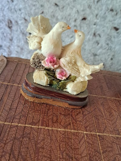 A figurine of two white birds, possibly doves, is placed on a wooden mat. The birds are depicted with orange beaks and small black eyes. One bird is slightly larger than the other and appears to be looking down at the smaller bird. They are positioned amongst pink and white flowers and green foliage, all mounted on a small, layered wooden base. The wooden mat is made of thin, closely spaced brown wooden slats, with some faint text or pattern printed on them. The background is a blurry, textured surface that appears to be light-colored and soft, possibly a carpet or fabric. The lighting suggests an indoor setting, and there are no indicators of a specific time of day or weather conditions. The overall impression is that of a decorative item. The faint text on the wooden mat is illegible.