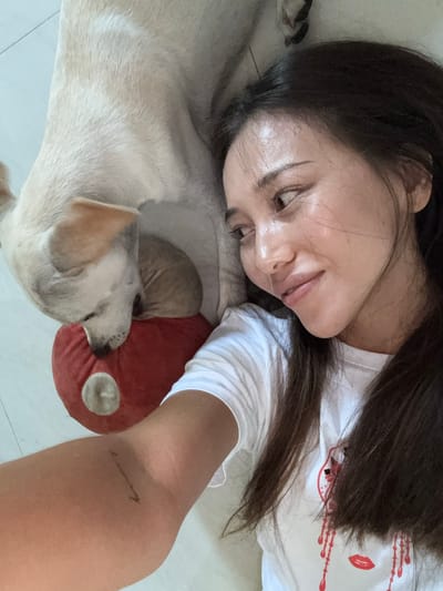 A young woman and a light-colored dog are lying on a white tiled floor. The dog is resting its head on a red stuffed toy. The woman is looking to the right of the frame with a slight smile. Her dark hair is partially obscuring her face and chest. She is wearing a white t-shirt with a red graphic design. The dog's fur is light brown and white. The environment appears to be indoors, likely a home in Baan Nok Sai, Thailand, given the context.