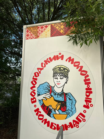 This image shows a close-up view of a logo on a white trailer, possibly a food truck or delivery vehicle, parked outdoors. The logo features a circular design with red Cyrillic text bordering a drawing of a young woman in traditional clothing. She wears a patterned hat and a blue polka-dot dress with red trim. Her eyes are closed, and she is pouring liquid from a yellow jug into a yellow bowl. Above the circular logo, the trailer has a decorative band with a pattern of red, yellow, and white geometric shapes. The background consists of lush green tree foliage and a blue sky, suggesting a daytime scene in an outdoor urban or suburban environment. The specific location in the city of Рубцово, Russia, is not visually discernible from the image itself.