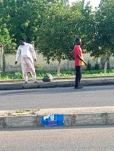 Two men stand on the side of a paved road in Garko, Nigeria. The man on the left wears a flowing light pink garment and white sneakers. The man on the right wears a red and yellow short-sleeved shirt and black trousers. Behind them, a line of trees with lush green foliage runs parallel to the road. In the foreground, a concrete curb separates the road from a patch of green vegetation. A dark green bag rests on the curb between the two men. A blue poster with white lettering and a white hand symbol is affixed to the curb in the lower right portion of the image. The scene appears to be outdoors during daylight.