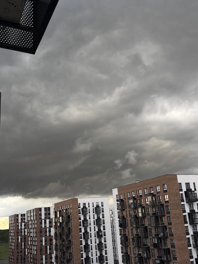 This image captures a view of apartment buildings against a dramatic, overcast sky in Misaylovo, Russia. The foreground features a dark, perforated structure, possibly part of a balcony or external walkway, extending from the top left corner. Below this, the sky is filled with heavy, dark grey clouds, suggesting impending rain or stormy weather. The middle ground is dominated by a row of modern apartment buildings with a repeating pattern of brown brick and white sections, punctuated by numerous dark-framed windows and balconies. The buildings are tall and appear to be densely packed. In the distance, on the left side of the frame, a strip of green landscape with trees can be seen, indicating a transition to a more natural or less urbanized area. The lighting is dim and diffused due to the cloud cover. No people are visible in the image.