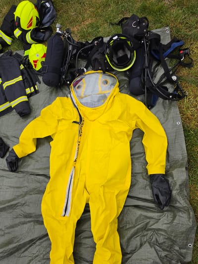 The image shows various pieces of emergency response equipment laid out on a gray tarp, possibly for inspection or inventory, against a backdrop of grass. The equipment includes a bright yellow hazmat suit with black gloves, a self-contained breathing apparatus (SCBA), fire helmets, and firefighter turnout gear. The scene is situated outdoors. The image was likely taken in Mlčechvosty, Czechia, as the location was provided.
