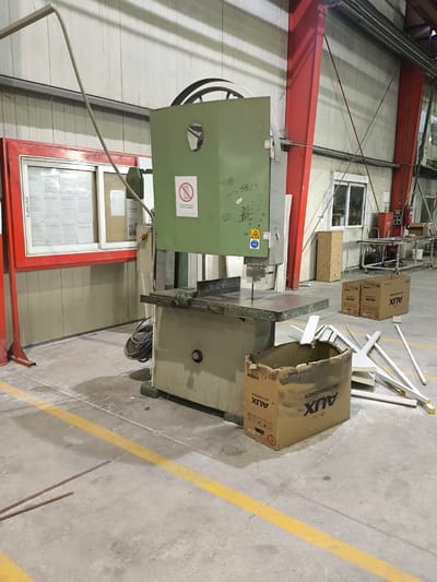 The image depicts an interior view of a factory or workshop in the Municipal Unit of Inofyta, Greece. The primary focus is a large, industrial band saw machine, painted a light green and beige, situated in the center of the frame. The machine has a prominent cutting blade and a sturdy metal table. To the left of the band saw, a red-framed display case with a glass front is visible, containing various documents and notices. A grey corrugated pipe extends from the upper left of the scene towards the band saw.

In the foreground, a cardboard box labeled "AUX AIR CONDITIONER" is placed on the floor. Scattered around the box are several white, elongated objects, possibly parts or materials. The floor is concrete, marked with a prominent yellow line, indicating a walkway or safety zone.

The background reveals the industrial environment with metallic support structures, possibly red beams, and corrugated metal walls. A window with a view of the outside is also present in the background, as are other pieces of industrial equipment and furniture, including a metal cart and a fire extinguisher. The lighting suggests an indoor setting with artificial illumination. There are no people visible in the image. The scene appears to be a static record of the workshop's layout and contents at the time the photograph was taken.