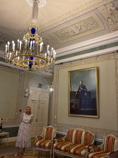 This image captures an interior scene within the Constantine Palace in Strelna, Russia. A woman, wearing a long, floral-patterned dress and light-colored shoes, stands in the foreground to the left of the frame. Her blonde hair is pulled back, and she is looking towards the viewer with her arms behind her back.
The room is opulently decorated, featuring a large, ornate golden chandelier hanging from a highly detailed ceiling adorned with plasterwork. To the right, a large, framed portrait of a man in a military uniform is displayed on the wall. In front of the portrait, a set of upholstered chairs and a sofa with striped upholstery in orange, cream, and gold are arranged. The walls are painted a pale green and cream, with decorative borders.
In the background, a white double door is visible, with an exit sign above it. Another smaller, decorative chandelier hangs to the right of the main chandelier. The floor is a polished wood parquet. The overall impression is one of a grand and historically significant interior.