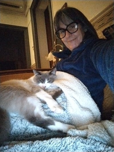 The scene depicts an indoor environment, likely a residence in Montélimar, France. A female subject occupies the right side of the frame, facing the camera with a slight smile. She has dark, shoulder-length hair, wears black-rimmed glasses, and is dressed in a dark blue, possibly fleece, long-sleeved garment. Adjacent to her on the left is a bi-color cat, primarily grey with white markings on its face, chest, and paws. The cat is lying down with its eyes partially closed, resting on a light blue, textured, fluffy blanket. A white, textured blanket, possibly a sherpa-style fabric, partially covers the cat and extends towards the human subject. In the background, a dark doorway or archway is visible on the left, along with a reflective surface and an ornamental light fixture featuring a yellowish, translucent shade. The overall lighting suggests an interior setting.