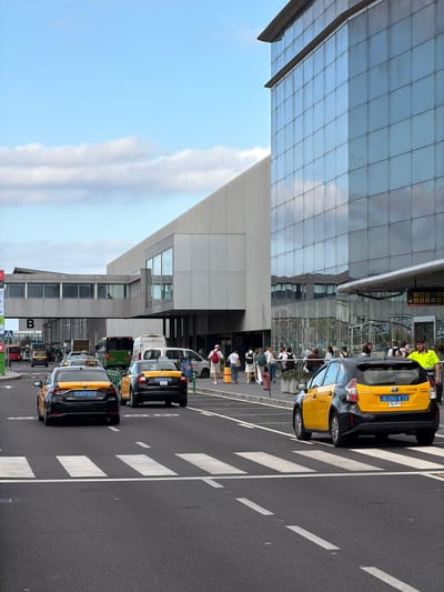 This is an outdoor, eye-level, vertical shot of an airport terminal building in El Prat De Llobregat, Spain. The terminal has a modern facade of glass panels that reflect the sky and clouds. To the left of the building, a pedestrian bridge connects to another section of the terminal. In front of the terminal, there is a road with several yellow and black taxis waiting or driving. Pedestrians are walking on the sidewalk next to the road. The road has a crosswalk. The sky is blue with scattered clouds. The scene appears to be during daytime.
