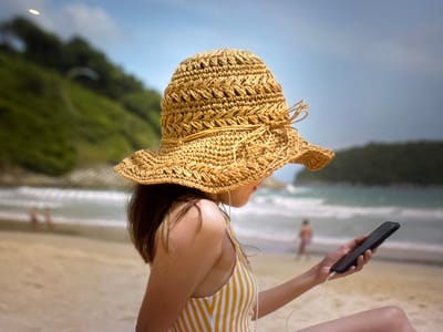 A woman wearing a straw hat and a yellow and white striped swimsuit sits on a beach, holding a black smartphone connected to white earbuds. The woman is viewed from the side, with her face obscured by the hat. In the background, the ocean meets the shore with gentle waves, and a green hillside rises behind the beach. Two blurry figures are visible in the distance on the beach and in the water. The sky above is a clear blue. The scene appears to be a relaxed vacation setting, typical of a coastal area. There is no indication that this scene is located in the city of Bevern, Germany.