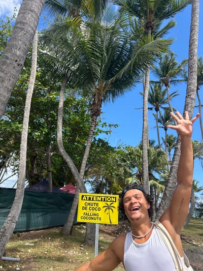 The image shows a man in Sainte-Anne, France, standing next to a sign that reads "ATTENTION Chute de Coco Falling Coconuts." He is fair-skinned, wearing a white tank top and a string of pearls, with dark hair and a black headband. His right arm is extended with his fingers splayed. He is smiling broadly and facing the camera. The sign is yellow with black lettering and a stylized image of a palm tree with coconuts. Behind the man and the sign are several tall palm trees with green fronds set against a clear blue sky. Green foliage is also visible in the background. There is a dark green covering in the background. The image is slightly rotated.