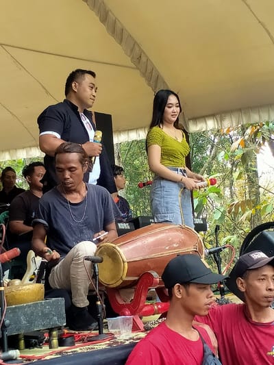 An outdoor musical performance is underway in Tamansari, Indonesia, beneath a light-colored tent canopy. A male singer/MC stands prominently on the left, facing right in profile, holding a gold-colored microphone. He wears a dark short-sleeved collared shirt over a white t-shirt. To his right, a female singer stands, facing slightly left and looking down, holding a microphone with red and yellow accents. She wears a light green ruched top and light blue denim jeans. In the immediate foreground, a male musician is seated, playing a brown wooden kendang (drum) mounted on a red stand. He wears a dark blue t-shirt and light-colored pants, holding a white cigarette in his right hand, with a silver chain around his neck. To his left, a small, brass-colored gong-like instrument rests on a black surface. Several other individuals are visible in the background and foreground, including musicians with Korg keyboards and crew members. The stage area is covered with a patterned mat, and audio equipment, including speakers and cables, is present. Two men in red t-shirts and baseball caps are partially visible in the bottom right foreground. Lush green trees and foliage are visible behind the tent, indicating a daytime outdoor event.