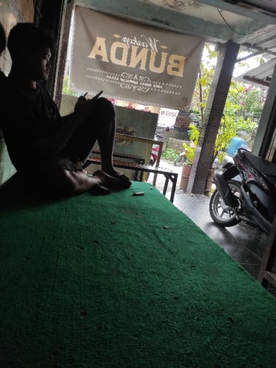 A young man, wearing a dark shirt, sits on a green artificial turf surface. The man is looking down at a cell phone or small device in his hand. The man appears to be in a storefront, with a sign reading "BUNDA" in gold lettering, above the entrance. There is a wooden table and chairs to the left of the man. To the right, outside the storefront, is a black motorcycle with a visible rear wheel. The street beyond the motorcycle is partially visible. The scene is located in Kecamatan Peterongan, Indonesia, at the coordinates -7.545 latitude and 112.277 longitude. There is a wooden door to the right of the sign. The door is open, and there is a glimpse of a dark-haired man, possibly entering or leaving the storefront. The doorway is shaded, suggesting the time is likely either morning or evening. A large green bush is visible behind the sign.