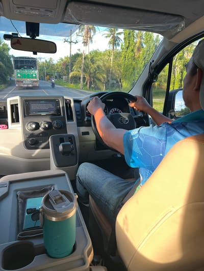 This media file shows a person driving a vehicle. The driver, wearing a blue and white patterned shirt and blue jeans, is seen from the chest up, with their hands on the steering wheel. The interior of the vehicle is visible, including the dashboard with a touchscreen display and various controls. A teal insulated cup is placed in a cup holder. Outside the vehicle, palm trees and greenery suggest a tropical or subtropical location. The sun is shining, casting a warm light on the scene, indicating it is daytime. There is a truck visible ahead on the road. The overall atmosphere appears to be a scenic drive on a sunny day.
Visible text includes "TOYOTA" on the steering wheel and a warning message on the touchscreen display that reads "CAUTION: Drive safely and obey traffic laws. You can help to prevent a serious or fatal accident by using safety features and restraints properly. Read safety and maintenance information in your Owner's Manual." There is also a "REAR" label visible on a control knob.