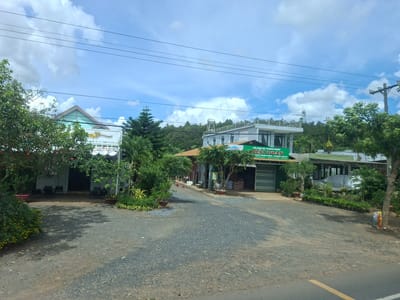 The image shows a commercial area with several buildings and lush greenery under a bright, cloudy sky. The scene appears to be in Phú An, Vietnam, as indicated by Vietnamese signage.
In the foreground, a gravelly dirt road leads into the commercial area. To the left, a building with a light-colored facade and a teal roof extension is visible, with outdoor seating consisting of chairs and small tables. A potted plant with yellow flowers is also situated in this area.
Further into the scene, a larger building with a white upper story and a green awning over the ground floor is prominent. A large green sign with white Vietnamese text and a logo hangs above the entrance. This building appears to be a storefront, possibly selling agricultural supplies, as suggested by the text on the sign and stacks of what look like bags of fertilizer inside. Several smaller potted plants are arranged in front of this building.
To the right of this building, another structure with a white upper floor and a darker roof is visible. This building has a balcony and several windows.
The background is characterized by dense trees and vegetation, with rolling hills beyond. Telephone poles and power lines are visible crossing the sky. The overall impression is of a rural or semi-rural commercial establishment in a tropical or subtropical climate. No people are visible in the image.