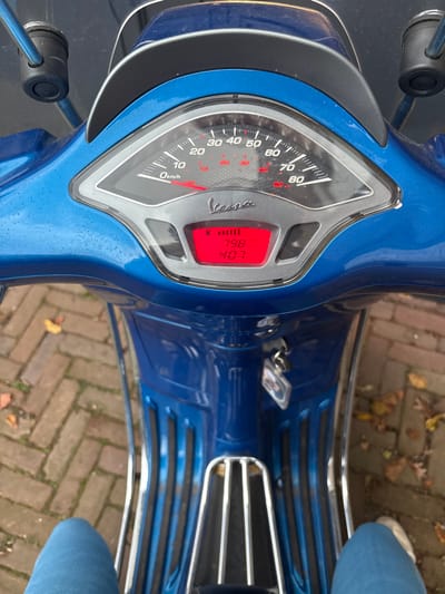 The image provides a first-person perspective from the seat of a stationary, blue metallic Vespa scooter, located in the city of Rosmalen, Netherlands. The scooter's handlebars and instrument cluster are central, with an analog speedometer visible, scaled from 0 to 80 km/h, indicating 0 km/h. Below the speedometer, a red digital display shows "798" and "407", likely representing odometer and trip meter readings. The Vespa brand logo is present on the dashboard. Keys with an attached keychain are inserted into the ignition. The scooter's surfaces exhibit small water droplets. The lower portions of a person's legs, wearing blue jeans and light-colored footwear, are visible at the bottom of the frame, indicating their position on the scooter. The ground beneath the scooter is comprised of reddish-brown brick paving, upon which several scattered dry leaves are visible.