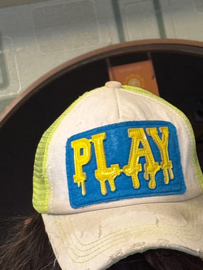 A close-up shot shows a partially visible white baseball cap resting on a dark surface. The cap features a blue rectangular patch with the word "PLAY" embroidered in yellow, vertically oriented. A thin band of green piping is visible along the brim's edge. The cap's fabric appears worn and slightly discolored. A small portion of dark hair is visible near the cap's edge. To the left, a portion of a brown shelf and a lighter-colored, textured surface are visible. There is no indication of a location in Novo-Nikol'skoe, Russia.