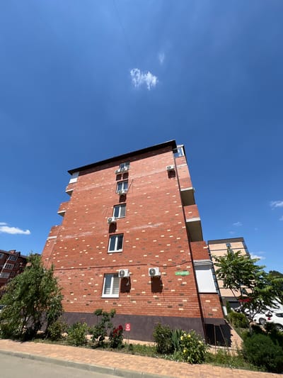 A brick apartment building is viewed from a low angle against a bright blue sky. The building has multiple windows and air conditioning units. There are trees and bushes in front of the building, and a paved walkway. In the background, another building is partially visible, along with parked cars. A street sign on the brick building reads "ул. Октябрьская 29". This suggests the location is Octyabrskaya Street, 29, in Novaya Adygeya, Russia.