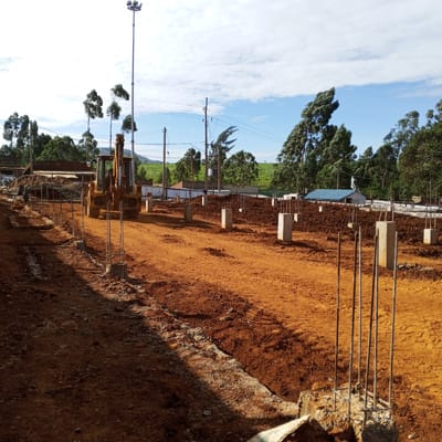 The image depicts a construction site in Borabu Chitago ward, Kenya. The ground is red earth, partially leveled. Several concrete pillars with rebar extending upwards are erected in a grid pattern. A yellow backhoe is parked on the left side of the frame. In the background, trees and buildings are visible, along with utility poles and wires. The sky is partly cloudy.