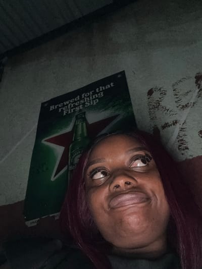 The image shows a close-up of a young Black woman with purple hair looking upwards with a playful expression. Her eyes are wide and her mouth is slightly open. In the background, a Heineken beer poster is visible, featuring a green bottle and a red star. The poster appears to be affixed to a textured, light-colored wall. The lighting is dim, casting shadows and creating a somewhat moody atmosphere. The scene appears to be indoors, possibly in a casual setting. There are no visible indicators of the specific location within Abuja, Nigeria, beyond the poster itself which is a global brand.