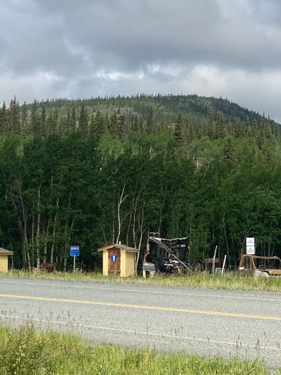 The image depicts a roadside scene in Haines Junction, Canada. In the foreground, a gray asphalt road with yellow double lines runs horizontally across the frame. The area adjacent to the road is covered with green grass and some wildflowers.
Behind the road, a line of trees forms a dense forest, with deciduous trees closer to the foreground and evergreen trees extending up a sloped hill in the background. The sky above is overcast with gray clouds.
On the right side of the road, there are several objects. A small, yellow wooden structure resembling an outhouse is visible. Next to it, a partially burned RV or mobile home is present, with its roof and interior structure exposed and charred. Further to the right, the remains of a vehicle, possibly a truck, are also visible, appearing burned and rusted. A white sign with black lettering is posted near the burned vehicle.
On the left side of the road, another small, yellow building is partially visible. A blue sign with white symbols is also present in the area between the two yellow buildings. The overall scene suggests an area with basic facilities, but also evidence of recent damage or disuse.