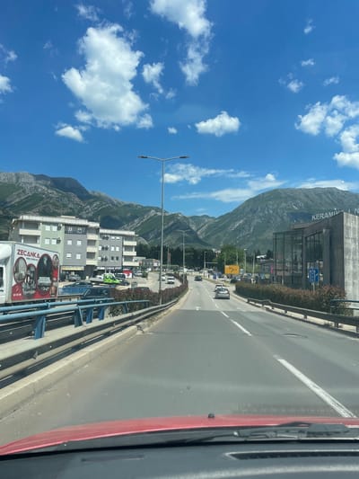 The image depicts a road in Bjeliši, Montenegro, as seen from a moving vehicle. A mountain range dominates the background under a blue sky with scattered clouds. A building with the word "KERAMIKA" visible on its facade stands on the right side of the road. Cars and street lights line the street, and a bridge with a blue railing is visible in the foreground. The side of a white truck, bearing the logo of "ZECANKA," is also present.