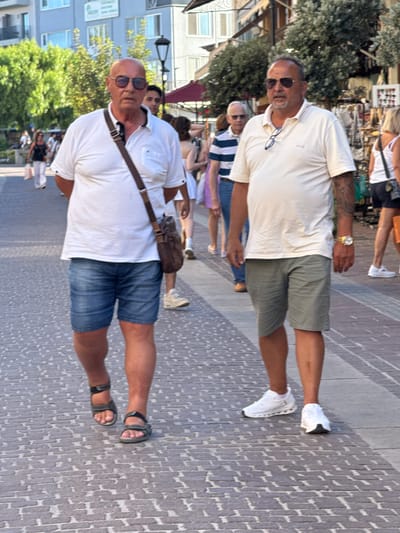 Two adult males are walking on a cobblestone pedestrian street in Chania, Greece. The man on the left is wearing a white polo shirt, blue denim shorts, and dark sandals. He has a brown satchel slung across his body and is wearing sunglasses. The man on the right is wearing a light-colored polo shirt, green shorts, and white athletic shoes. He has sunglasses resting on his shirt collar and is wearing a gold wristwatch. Several other pedestrians are visible in the background, walking in the same direction. Buildings line the street, with shops and trees on either side. The lighting suggests it is daytime, possibly late afternoon, with the sun casting long shadows.