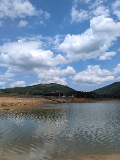 The image depicts a reservoir or lake located near the city of Ngoc Lac Commune, Vietnam. The foreground is dominated by the calm surface of the water, reflecting the sky and surrounding landscape. The shore is lined with rocks and a sloped, textured embankment, possibly part of a dam structure. In the mid-ground, a metallic walkway or pier extends into the water, with some blue structures and pipes visible on it. Beyond the water and embankment, rolling green hills covered in vegetation rise towards the horizon. The sky is a vibrant blue, dotted with fluffy white clouds. The scene appears to be a natural landscape with some man-made infrastructure. There are no people or animals visible in the image.