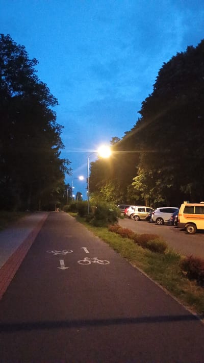 The image depicts a bicycle path at dusk in Sokolov, Czechia. The sky is a deep blue, transitioning from twilight to night, with a few scattered clouds visible. The path is paved and has painted bicycle lane markers, including arrows and symbols of bicycles. A red-brick border runs along one edge of the path, separating it from a grassy area with some bushes and trees.
Along the side of the path, several vehicles are parked, including what appears to be an ambulance or a similar service vehicle, along with at least two smaller cars. These vehicles are illuminated by streetlights.
Several streetlights are visible along the path, casting a warm glow and illuminating the immediate surroundings. The trees lining the path are dark silhouettes against the twilight sky. The overall mood of the image is peaceful and quiet, suggesting the end of a day. No people are visible in the image. The text "i" is visible next to a bicycle symbol on the path.