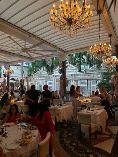The image depicts an upscale outdoor dining area at The Villa Casa Casuarina, formerly the Versace Mansion, located on Ocean Drive in Miami Beach, United States.
The scene is set under a large, retractable or permanent light-colored awning ceiling, from which multiple ornate crystal chandeliers hang, casting a warm, golden light. A large, industrial-style ceiling fan is also visible. The ground is covered with a patterned mosaic tile floor.
In the background, the distinctive classical architecture of the Versace Mansion is prominent, featuring white stucco walls adorned with intricate relief work, pilasters, and friezes, consistent with the "Thousand Mosaic Pool" area. Lush tropical foliage, including tall palm-like trees, rises above the mansion walls. A large decorative urn is visible near a circular tower structure to the left. A wall-mounted fan is affixed to a column.
The dining area is furnished with numerous square and rectangular tables, draped with white tablecloths. Each table is set with white plates, glasses, and cutlery. Several tables feature distinctive menus with gold and black patterns. Upholstered dining chairs, primarily light in color, are arranged around the tables.
Approximately twenty to thirty individuals are present, engaging in various social activities. In the foreground, two women are seated at a table. One, on the far left, wears a red top and looks to her left, with a prominent tattoo on her left arm. Another woman in a red top/dress is partially obscured, reaching or gesturing towards the table. In the mid-ground, a man in a black shirt stands with his back to the viewer, conversing with another man in a black polo shirt and eyeglasses. Towards the right, groups of people stand and sit around tables; a woman in a black dress walks away from the camera, and another in a striped dress stands near a table. A person is partially visible in the water of a pool in the mid-right background, behind some diners. The overall attire suggests evening wear ranging from casual to semi-formal. The lighting indicates dusk or evening hours, with artificial illumination creating the primary light source.
