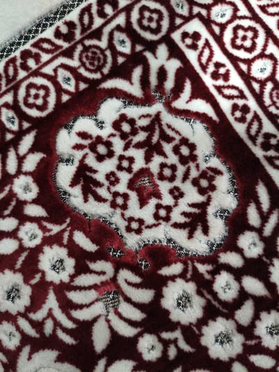 This is a close-up, overhead shot of a maroon and white rug. The rug features a repeating floral pattern with small, silver-threaded accents in the center of some of the flowers. The pattern is intricate, with curvilinear motifs and geometric elements. The rug appears to be made of a soft, plush material. The lighting is even, suggesting an indoor setting. There is no visible text. The image does not contain any people, and there is no discernible activity or event taking place. The specific location of Keudematangkuli, Indonesia, is provided as context but is not visually evident in the image itself.