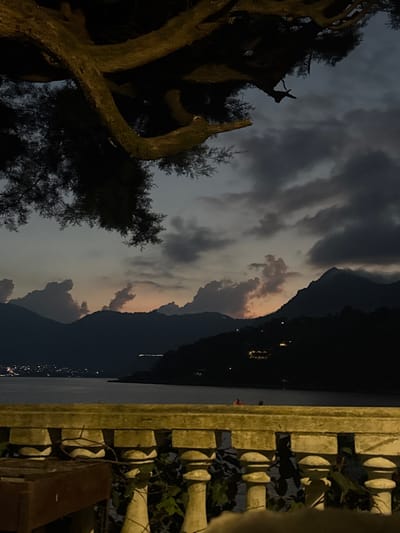 A twilight view features a tree with dark foliage on the left. In the center, mountains are framed by illuminated clouds with a dark sky above. The right edge of the frame shows a concrete railing with foliage growing around it. The overall impression is a serene landscape captured in the fading light.