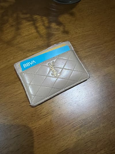 The image shows a beige, quilted leather cardholder with a gold YSL logo. A blue BBVA bank card is partially inserted into one of the card slots. The cardholder rests on a wooden surface. The location is unknown and there are no people present. It is unknown if the item or individual can be found in Villahermosa, Mexico.
