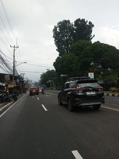 This image depicts a street scene captured on an overcast day in Dawathang_Dorjibi_ Kashingtsawa, Bhutan. The perspective is from a low angle on the asphalt road, looking forward down a multi-lane thoroughfare.

The road features white dashed lane markings, indicating at least two lanes of traffic moving away from the viewer. A black Daihatsu Terios/Toyota Rush SUV with a roof rack is in the rightmost lane, displaying the license plate "A 3590 UMC" on its rear. Ahead of it, a red sedan proceeds in the same lane. To the left of the red sedan, a single individual rides a white scooter or motorcycle, also moving away from the camera. Several motorcycles are parked at an angle on the left side of the road, adjacent to a concrete curb.

The left side of the scene is lined with low-rise buildings exhibiting worn facades, some with corrugated metal awnings. A prominent yellow rectangular sign on one building reads "SERUASLI". Multiple utility poles rise from the left, heavily laden with a dense network of black overhead wires.

On the right side of the road, a concrete curb separates the roadway from a landscaped median strip featuring short green shrubs and a substantial treeline composed of tall, mature trees with dark green foliage. A yellow and blue rectangular sign, partially obscured by trees, displays the letters "PLN". In the distant background, hazy mountains or hills are visible beneath a uniformly grey, cloudy sky. No other discernible human figures are present apart from the scooter rider.