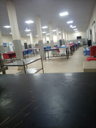 A long, bright cafeteria-style room is shown. Tables and chairs in blue and red are arranged in rows. White pillars line the room, supporting a ceiling with recessed lights. A metal cart is visible on the left. A dark surface occupies the foreground. A water cooler and other equipment are visible on the right.