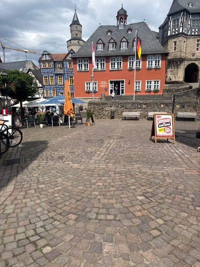 This image captures a vibrant street scene in Idstein, Germany, featuring a cobblestone square surrounded by historic architecture. In the foreground, a detailed cobblestone ground leads to an outdoor seating area with tables, chairs, and large umbrellas, where a few people are present. A bicycle is parked near the foreground on the right.

The background is dominated by several distinct buildings. On the left, a traditional half-timbered building with dark wood and white plaster contrasts with a striking red building featuring multiple white-framed windows, some adorned with flower boxes. A German flag (black, red, and gold) flies from a pole in front of the red building. Adjacent to it, a building displays a unique blue and yellow geometric pattern on its facade. Further left, a stone tower with a spire rises against a cloudy sky, and a construction crane is partially visible in the distance.

A prominent wooden sandwich board sign in the middle of the square announces a sale, with visible German text: "AUSVERKAUF GESCHÄFTSAUFGABE PREISNACHLASS 50% vollwertig regional," indicating a "clearance sale, business closure, 50% discount." Another sign partially visible on the red building reads "Rathaus der Stadt [Idstein]," confirming it as the town hall. The scene suggests a daytime setting, possibly under overcast skies, with a relaxed atmosphere typical of a European town square.