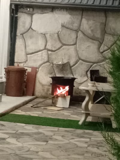 In Sasbaga, Uzbekistan, an outdoor area features a stone-patterned wall and a tiled floor. A small, dark-colored stove with a fire burning inside is centrally placed. To the left of the stove, a rustic brown metal container with handles and a lid stands. Adjacent to it is a reddish rectangular object. A light-colored table with decorative legs is situated to the right of the stove, holding a white bowl. A portion of a green plant is visible in the foreground, on the right side of the image.