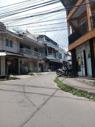 The image displays an urban street scene in Bagochen Boolingpang Ueling, Bhutan, under an overcast sky. A paved, slightly curved asphalt road, exhibiting signs of wear, occupies the foreground and midground. Buildings line both sides of the thoroughfare. On the left, a sequence of multi-story structures, primarily light-colored, features ground-level commercial fronts and upper-story residential sections with balconies and white balustrades. A satellite dish is visible on the roof of one building, and a red circular sign on a pole is positioned near the left curb. One visible sign on a shop reads "TOKO MORONOO." On the right, a distinct multi-story building with an orange-brown facade and black balcony railings features a ground-floor glass storefront displaying large posters depicting women's faces. Two dark-colored motorcycles are parked on the sidewalk adjacent to this building. Green foliage grows along the concrete curb on the right side of the road. A dense and complex network of black electrical and communication cables extends across the sky above the street and buildings. No individuals or specific human actions are clearly discernible within the frame.