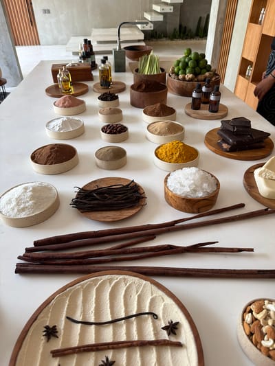 A large, rectangular white table is centered within a modern, minimalist interior in Tibubeneng, Indonesia, serving as a display surface for an array of natural ingredients and products.

The table is densely covered with various items arranged in ceramic and wooden bowls, and on circular wooden trays. From the bottom left, a wooden oval tray contains fine white powder raked into a pattern, adorned with star anise, small cinnamon pieces, and a vanilla bean. Above this, several long, dark reddish-brown cinnamon sticks are laid horizontally on the white table.

Moving upwards and right across the table:
Numerous light-colored ceramic bowls contain various powders: white (e.g., coconut powder, starch), reddish-brown, greenish-brown, light brown granular, and dark reddish-brown (e.g., cocoa/coffee grounds).
Several wooden circular trays hold different items: two small glass bottles with clear yellow liquid (oils), a pile of dark cocoa beans, a bundle of dark vanilla bean pods, and a stack of dark chocolate blocks.
Other wooden bowls contain specific ingredients: a large quantity of green limes with ginger roots at their base, bundles of green lemongrass stalks, and clear, irregularly shaped white crystals (sea salt or sugar).
Additional items include a ceramic bowl of vibrant yellow powder (turmeric) and another filled with a mix of almonds and cashews. A light-colored solid block (e.g., shea butter) rests on a wooden tray.
Further back, three dark glass bottles with brown liquid (essential oils) are visible on a wooden tray. A wooden tissue box and additional small bottles are also present.

A person's right arm and a portion of their torso, clothed in a dark garment with a light pattern, are visible on the far right side of the image, suggesting human presence near the display.

The background features light gray concrete walls. On the left, a white staircase with open risers ascends, with small green plants visible beneath it. A dark metal faucet or fixture is positioned in the mid-background. On the right, light-colored wooden shelving units containing small bottles are partially visible behind the individual. The lighting is bright and even throughout the scene.