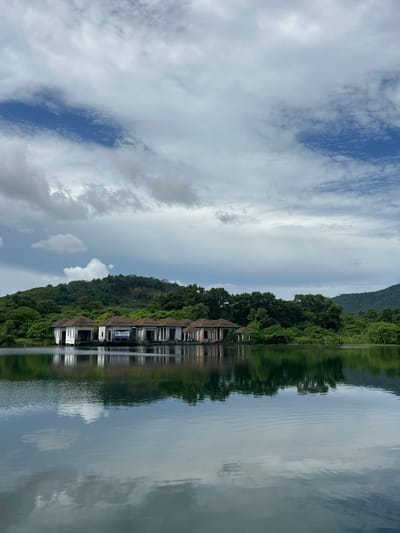 The image displays a serene waterside scene, likely in Si Sunthon, Thailand, under an overcast sky. The main subjects are a line of modern buildings nestled amongst lush green trees along the water's edge. The buildings appear to be holiday villas or residences, characterized by white facades and dark roofs, with multiple windows. A calm body of water stretches out in front of them, reflecting the sky, trees, and buildings, creating a symmetrical and tranquil atmosphere.
The time of day is difficult to determine precisely due to the cloudy conditions, but the soft light suggests it could be late morning or afternoon. The weather is cloudy, with scattered patches of blue sky visible, indicating a mix of sun and clouds. The abundant greenery and the style of the architecture are consistent with a tropical or subtropical location.
A notable detail is a sign visible on one of the buildings. It is rectangular and appears to have some text and graphics, though the details are not clear enough to read. The overall mood evoked by the image is one of peace and natural beauty, with the reflections in the water adding a sense of depth and calmness. There are no people visible in the frame, emphasizing the quiet and secluded nature of the location.