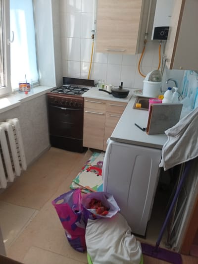 The image shows a messy, lived-in kitchen in Krymsk, Russia. A gas stove is positioned next to a white washing machine. The countertops are cluttered with various items, including a pot, bottles, and what appears to be a box. A window with a white curtain is visible on the left, and a radiator sits below it. On the floor, there are shopping bags and a child's play mat. A gas water heater is mounted on the tiled wall above the countertops. The overall impression is of a functional but unkempt living space.