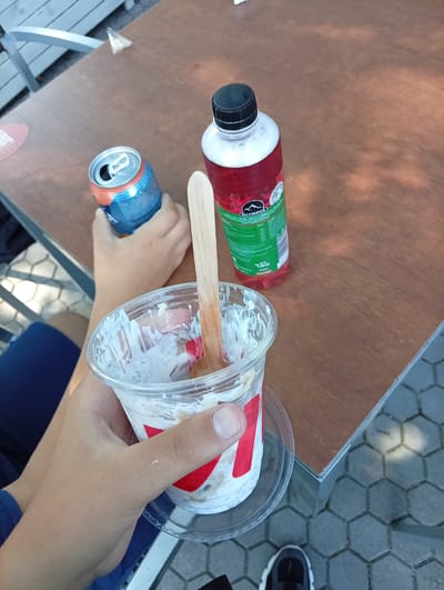 In Devin, Bulgaria, a person's hands are holding a can of soda and a cup of dessert on a wooden table. The can, mostly blue and orange, is held in the left hand, while the right hand grips a clear plastic cup containing a white, creamy dessert with a wooden spoon. A bottle of red liquid with a green label is also visible on the table. The background shows a textured gray surface and hints of outdoor seating.