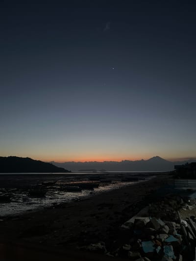 A twilight scene unfolds over a coastal landscape in Kertasari, Indonesia, featuring a beach, the sea, and distant landforms. The sky transitions from a deep indigo at the top to a soft gradient of blue and orange near the horizon, suggesting the period just before sunrise or after sunset. Two bright celestial bodies, likely planets or bright stars, are visible in the upper portion of the sky, along with a wispy cloud formation.
The foreground is dominated by a dark, sandy beach littered with rocks and debris. To the left, a muddy, exposed seabed is visible, reflecting the dim light and dotted with what appear to be remnants of structures or boats. A dark, silhouetted hill or island rises from the left side of the frame, its outline sharp against the fading sky.
Across the water, a hazy mountain range stretches horizontally, with a distinct, conical peak standing out on the right side. The water itself is calm, with the faint glow of the horizon reflecting on its surface. On the far right of the image, the dark silhouettes of buildings and construction materials are visible, indicating some level of human habitation or development near the coast. The overall atmosphere is serene and tranquil, characteristic of dawn or dusk. There are no people or animals visible in the frame.