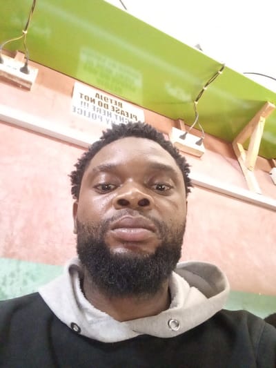 The image displays a headshot of a dark-complexioned male with a full black beard and short, dark, kinky hair, looking directly at the camera with a neutral expression. He is wearing a black hoodie with a grey hood lining and silver eyelets. The scene is indoors, located in Magboro, Nigeria. The background features a peach-colored wall with multiple white electrical outlets and exposed black wiring running along it. Above the subject's head, a white sign with black text is visible, appearing inverted and reflected, which, when deciphered, reads "PLEASE DO NOT RETAIL/RETAIN HERE !!! POLICE". Above this sign, a horizontal green panel or shelf extends across the upper portion of the frame, supported by a light-colored wooden bracket on the right. The area is brightly lit, likely by overhead artificial lighting.