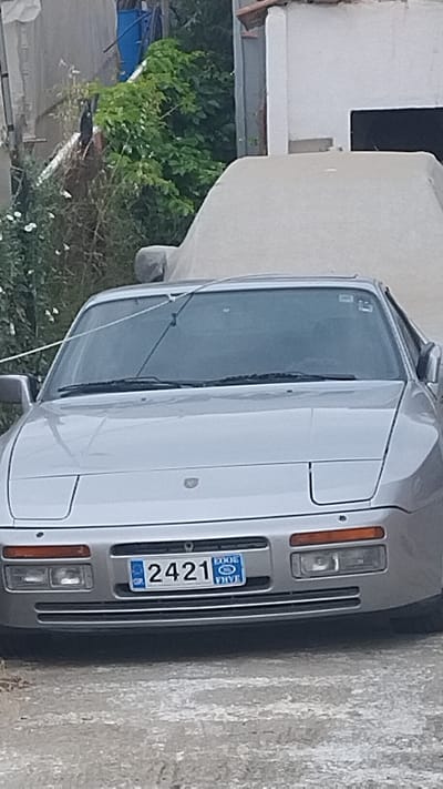 The image displays the front of a silver Porsche 944, parked outdoors in an urban or semi-urban setting, possibly in Νέα Κερασιά, Greece, given the context. The car's pop-up headlights are down. A blue and white license plate is visible on the front bumper, reading "2421". The car is partially covered by a light-colored tarp. In the background, there are dense green bushes and a white building with a dark opening, suggesting a residential or storage area. The ground in front of the car is paved with rough, grey material.