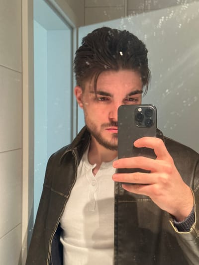 An individual is depicted in a mirror selfie taken within an interior space in Bertrange, Luxembourg. The subject is a male with dark, swept-back hair and a short beard. He is wearing a dark brown leather jacket over a white long-sleeved Henley shirt. His right hand holds a dark-colored smartphone, featuring a triple-lens camera array, positioned to capture his reflection. The subject's gaze is directed towards the camera. The background, visible through the reflection, consists of light-colored walls, possibly tiled, and a partial doorframe or architectural opening on the left. The mirror surface exhibits minor speckling or dust particles.