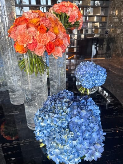 The scene depicts an interior setting, likely a commercial or public space, in Bawshar Province, Oman, showcasing multiple floral arrangements on a dark, reflective surface. In the foreground and midground, two prominent bouquets composed of orange, coral, and light pink roses, mixed with similarly colored carnations, are displayed. Each bouquet is contained within a tall, clear cylindrical glass vase filled with water, positioned vertically.
To the right and slightly lower, two arrangements of blue hydrangeas are present. The arrangement in the immediate foreground is a dense cluster of blue florets in a low, round, clear glass bowl, exhibiting varying shades of blue from light to medium. Behind it, another similar but slightly smaller arrangement of blue hydrangeas rests in an identical round glass bowl.
All floral displays are situated on a highly polished, dark black marble or granite surface, which exhibits subtle white veining and reflects the ambient light and portions of the flowers. The background features a wall composed of numerous rectangular mirrored or highly reflective panels, arranged in a grid-like pattern. These panels reflect distorted images of the environment, including architectural elements and diffuse light, suggesting an indoor, possibly upscale, setting. No individuals are visible in the image.