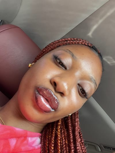 This image presents a close-up, angled portrait of a single individual, appearing female, positioned within the interior of a vehicle. The individual has dark brown skin, with dark eyes looking slightly upward towards the camera. Their eyebrows are dark and well-defined. The most prominent feature is a set of full lips coated in a dark, highly glossy product, formed into a pout. The hair is styled in small, reddish-brown box braids. Small gold-colored stud earrings are visible in both earlobes, and a thin gold-colored neck chain is present. A portion of a pink or red patterned garment is visible on the left. The background reveals the interior of a vehicle, including a dark reddish-brown padded surface, likely a seat or headrest, and a lighter-colored ceiling/headliner reflecting ambient light. A car air vent is partially visible in the bottom right. The specific geographical location of Ikota, Nigeria, cannot be determined from the visual information provided in the image.